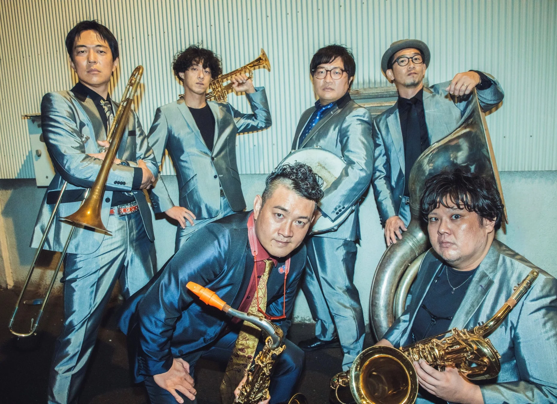 光沢のあるスーツを着た6人の男性が、トロンボーンやサックスなどの楽器を手にポーズをとっている