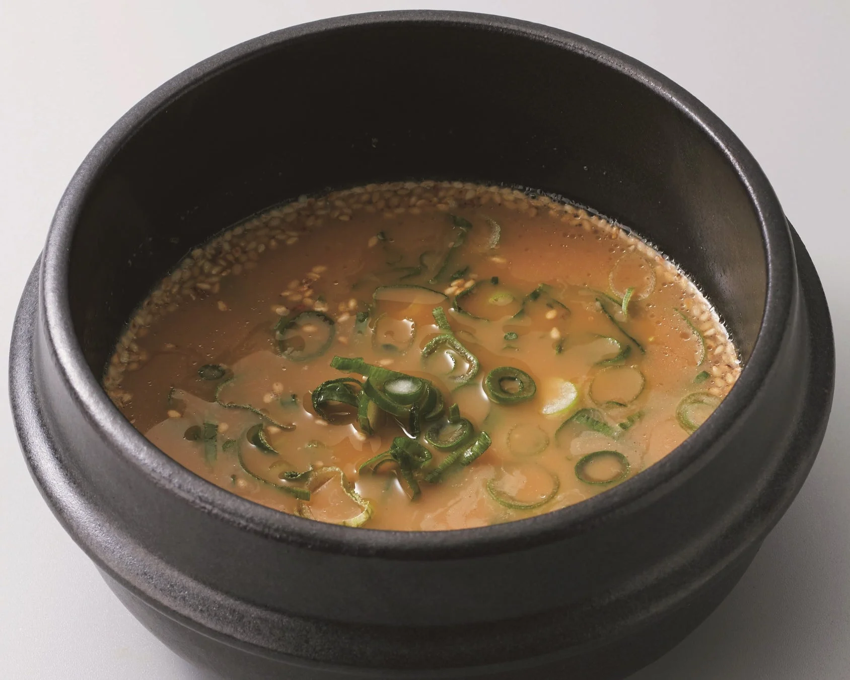 黒い陶器の器に入った温かいスープの写真です。淡い茶色のスープには、たっぷりの刻みネギと白いごまが散らされており、食欲をそそる一品です。