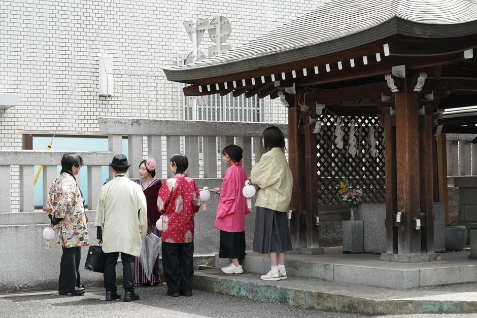 和装や現代的な服装の人々が、白いレンガの建物と伝統的な木造の社殿らしき建物の前で集まっている