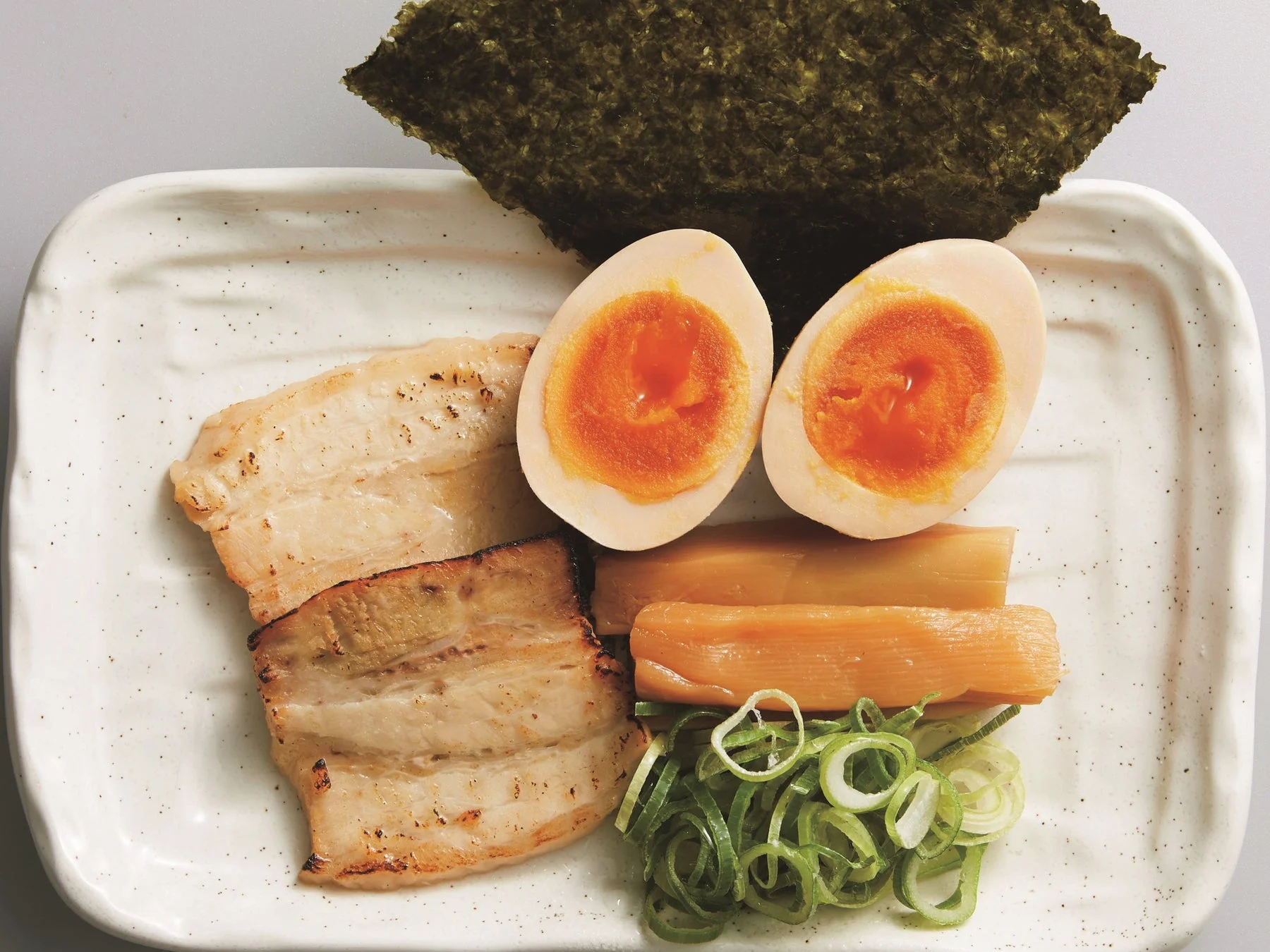 白い皿に盛られたラーメンの具材のセットです。香ばしく焼かれたチャーシュー、とろりとした半熟の味玉、メンマ、刻みネギ、そして海苔が美しく配置されています。