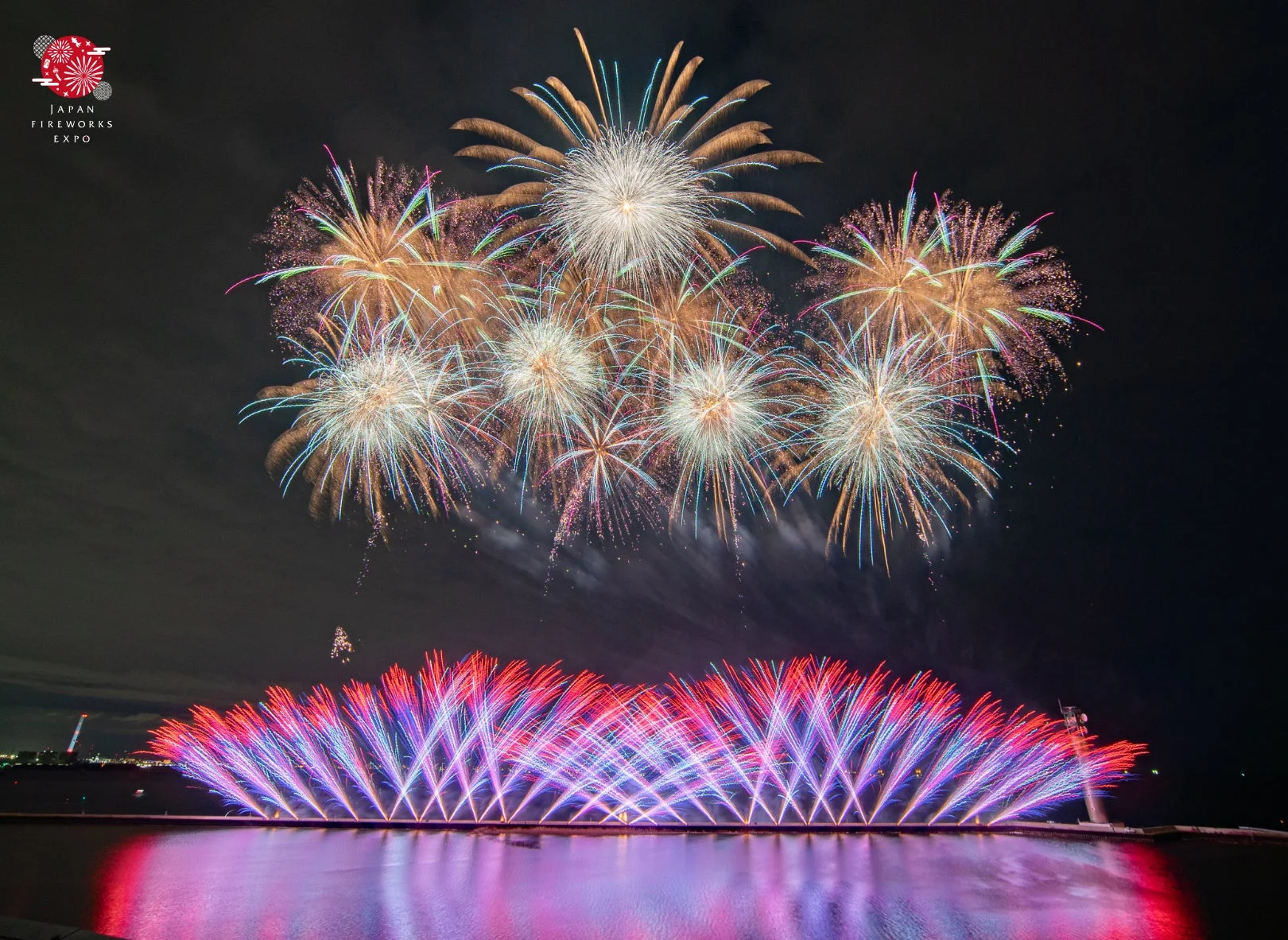 夜空に咲き誇る色とりどりの打ち上げ花火と、水面に映る赤と青の扇状の花火が美しい、壮大な花火大会の様子。