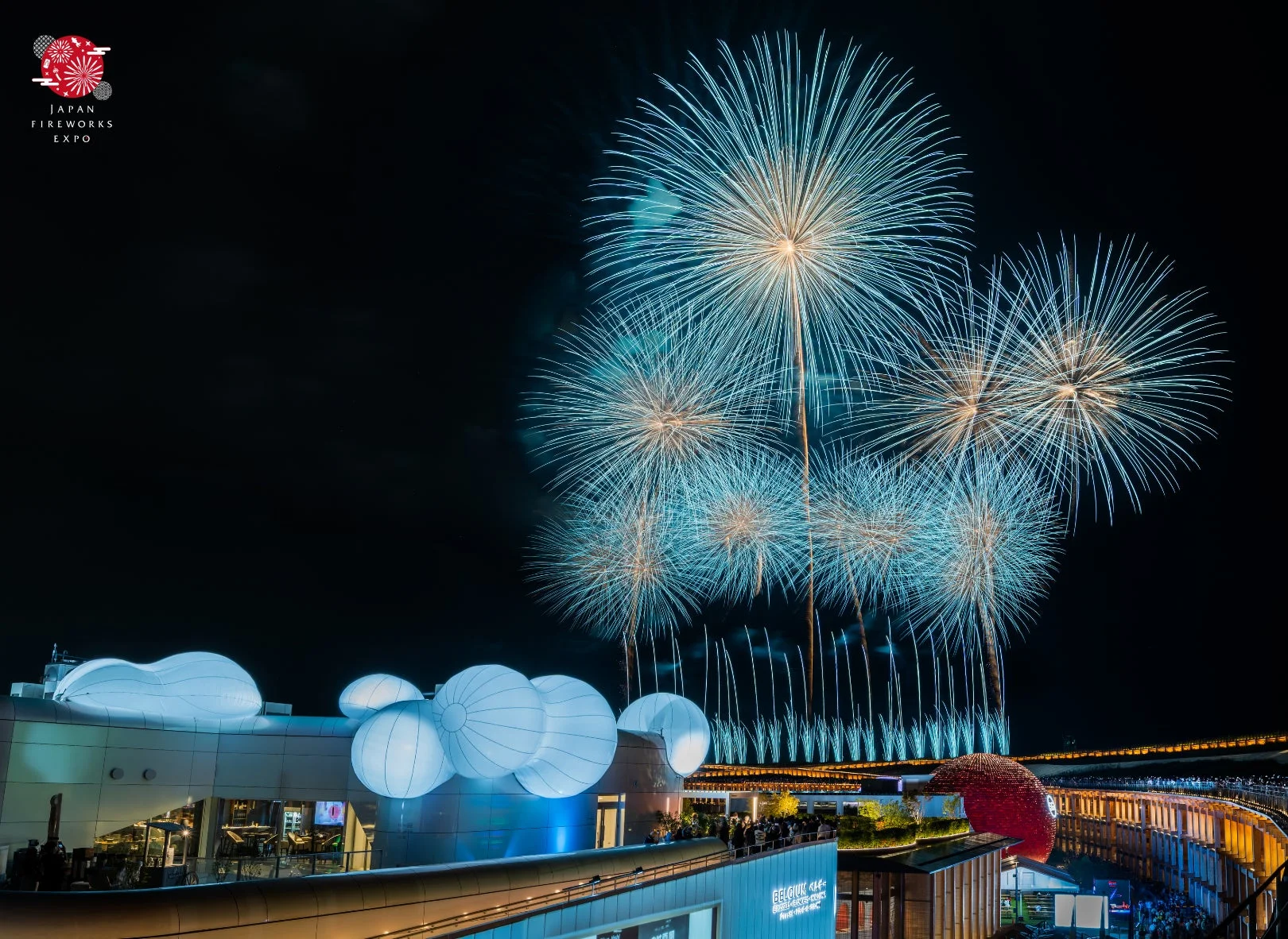 夜空に青と白の美しい花火が打ち上げられ、下にはモダンな建物がライトアップされています。