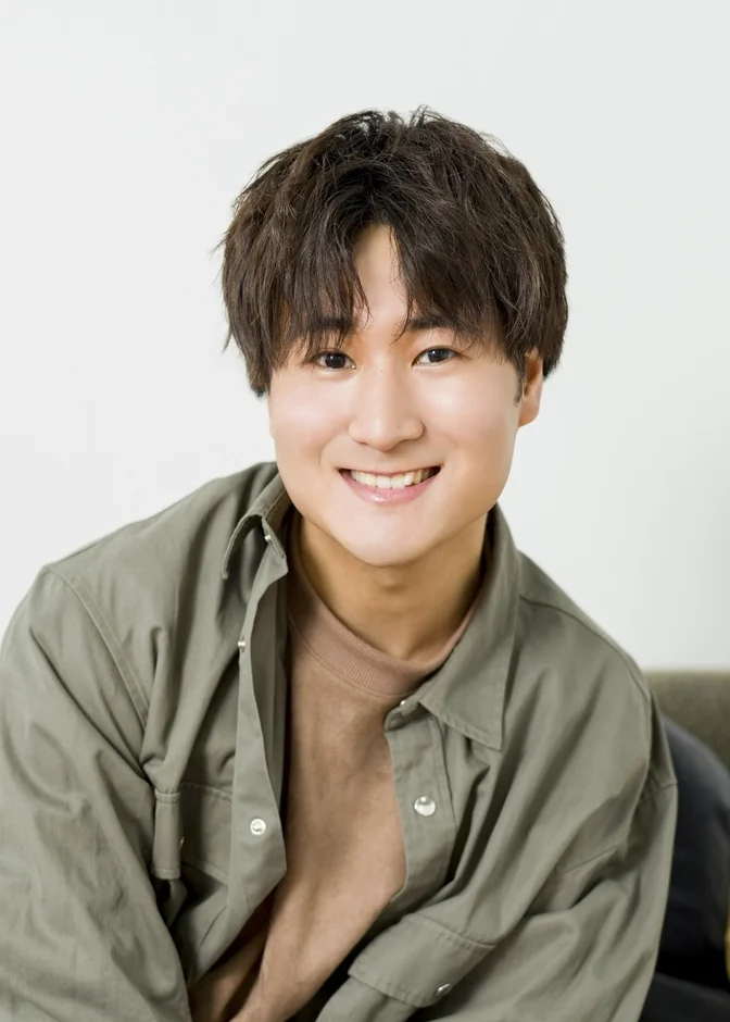 白い背景の前で、カーキ色のシャツを着た若い男性がカメラに向かって笑顔を見せているポートレート写真です。