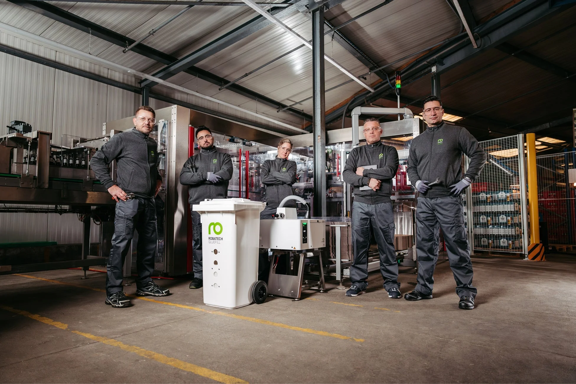 工場内で作業服と安全メガネを着用した5人の従業員が、機械設備の前で立ちポーズをとっています。彼らは「ROBATECH GLUEFILL」の製品やサービスに関わるチームのようです。