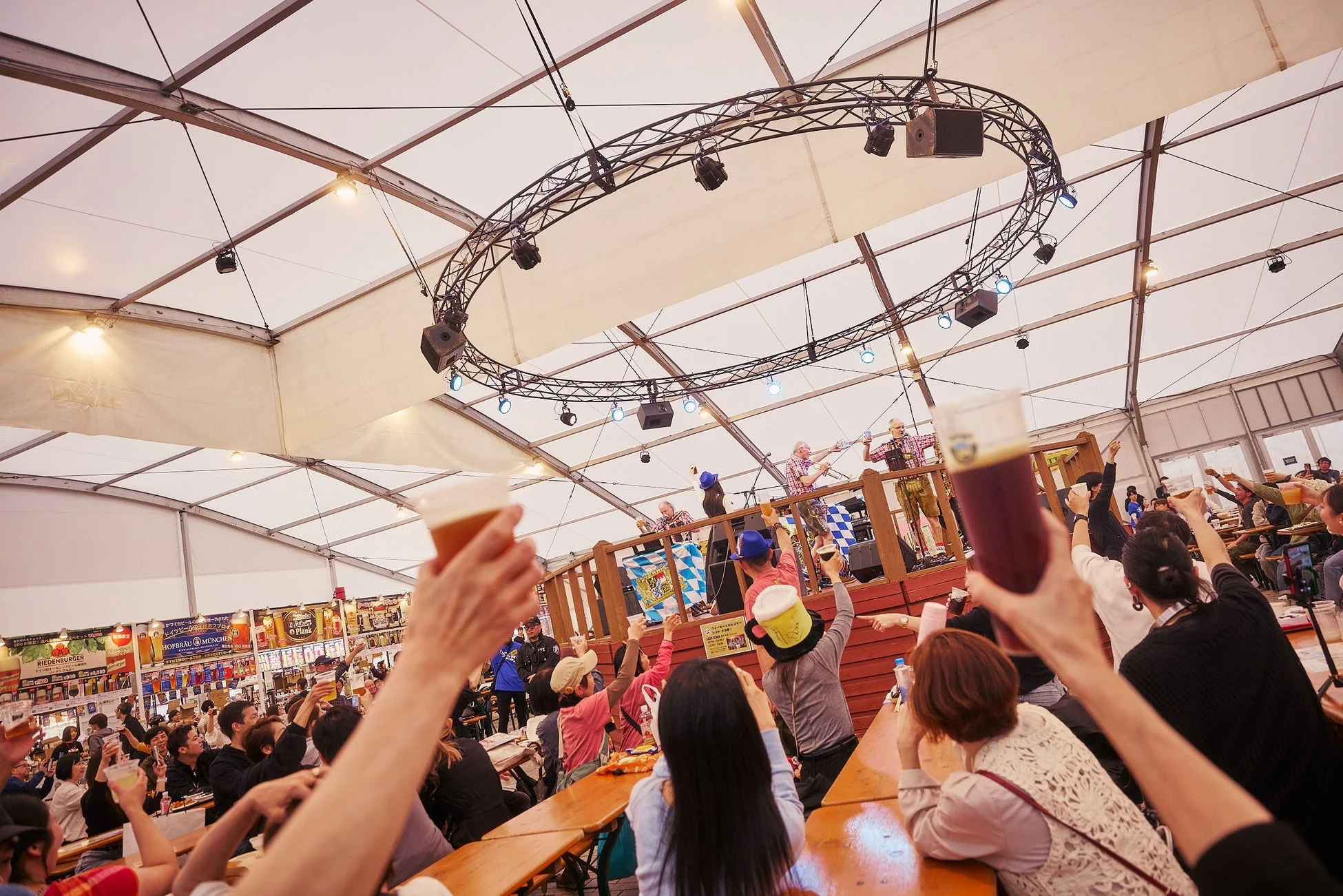 大きなテントの中で開催されているビール祭りやオクトーバーフェストのようなイベントの様子。多くの人々が木製のテーブルに座り、グラスを掲げて楽しんでおり、ステージでは伝統的な衣装を着たパフォーマーが盛り上げている