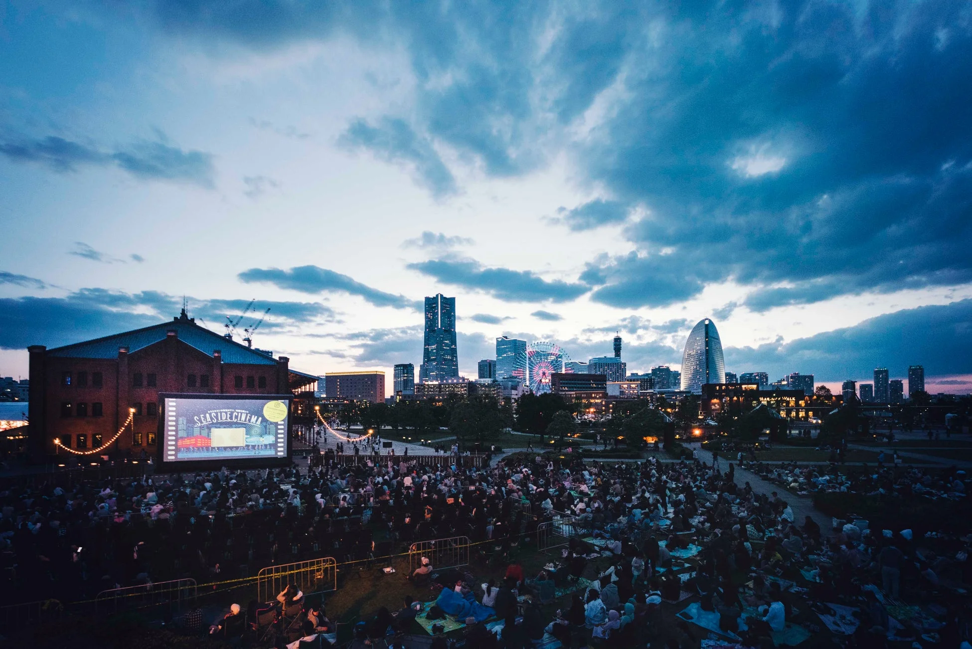 夕暮れの横浜赤レンガ倉庫を背景に、多くの人々が広場で野外映画「SEASIDE CINEMA」を楽しむ様子
