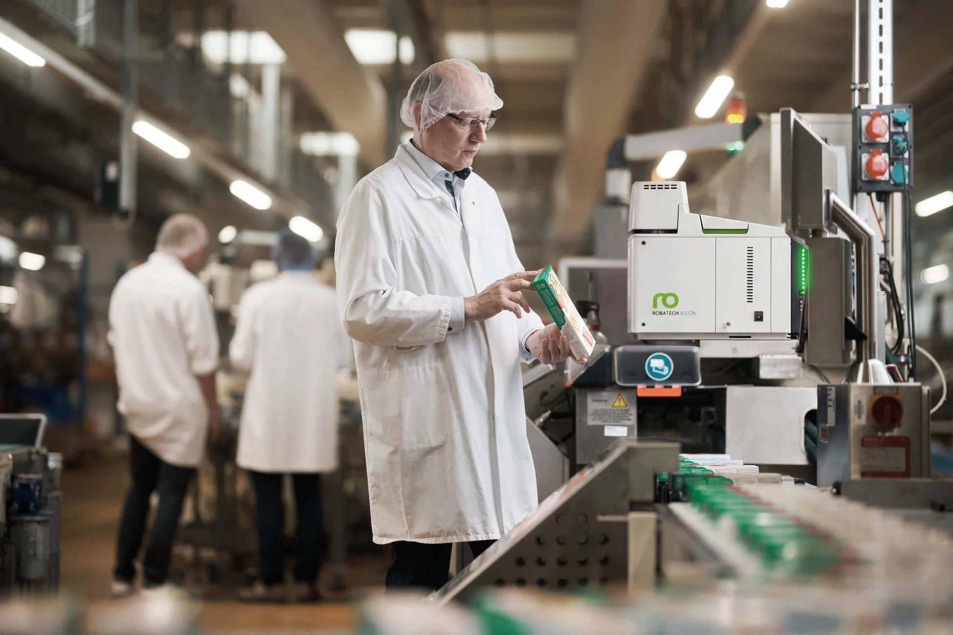 白衣を着た作業員が工場内の生産ラインで製品パッケージを検査している。ROBATECH VISIONの機械が設置され、製品の品質管理が行われている様子がうかがえる。