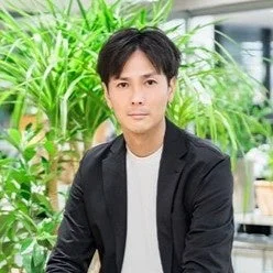 黒いジャケットと白いTシャツを着た男性がカメラを見つめています。背景には緑豊かな植物が配置されており、屋内での撮影であることが伺えます。