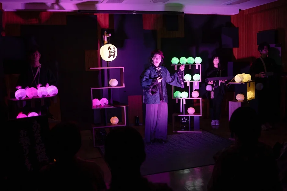暗い屋内で、提灯や光るオブジェが飾られた舞台で、数人の演者がパフォーマンスを行っている様子