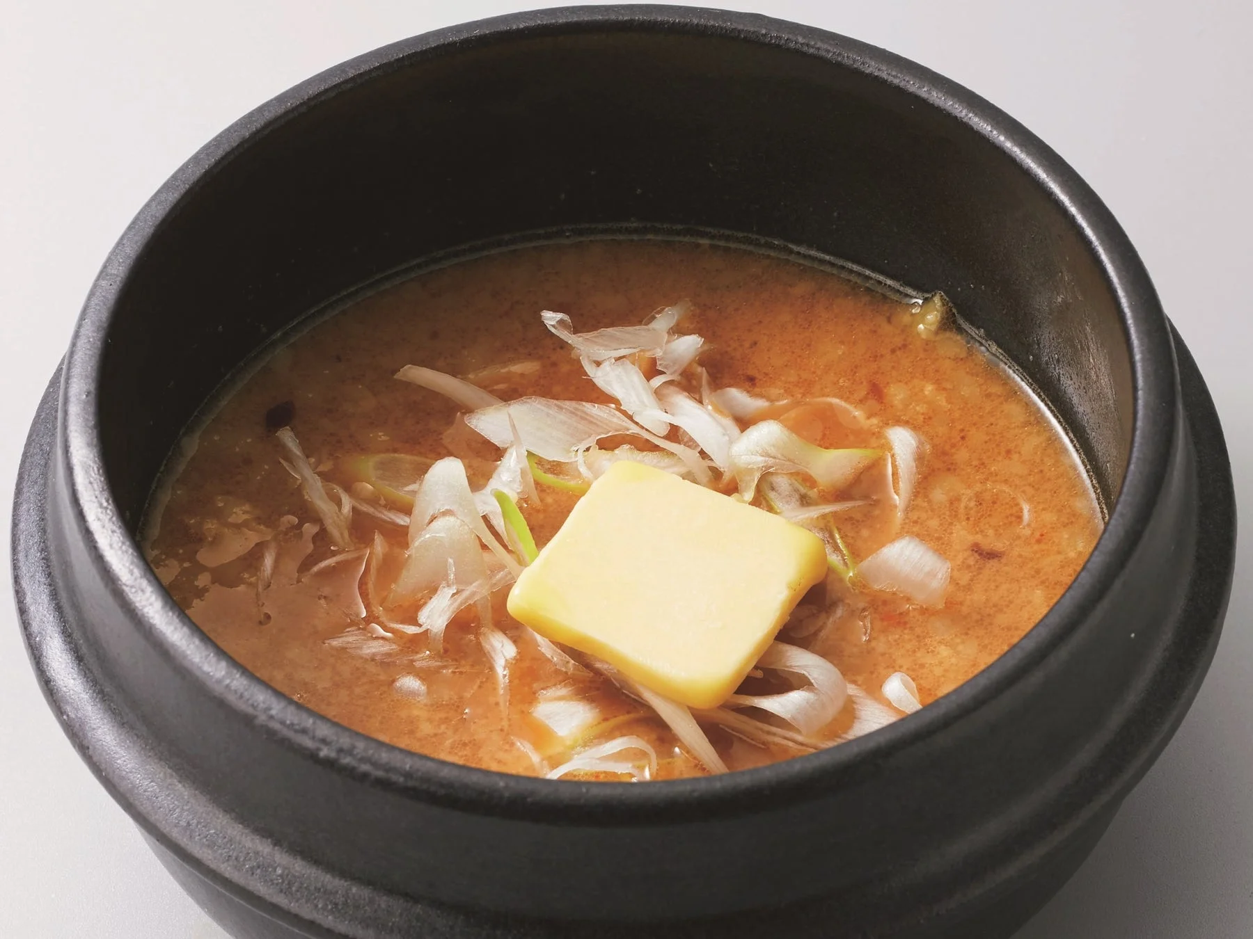 黒い器に入った、ネギとバターが乗った温かいスープの写真です。スープは赤みがかった茶色をしており、味噌ベースか辛味のある味付けを連想させます。