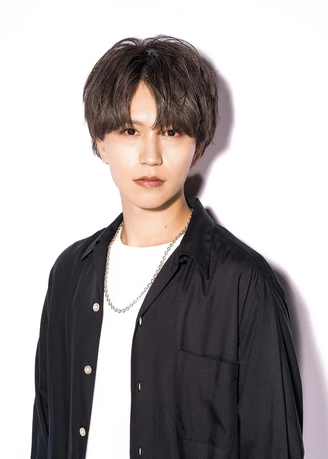 白い背景の前で、黒い開襟シャツと白いTシャツ、シルバーのネックレスを身につけた若い男性のポートレートです。