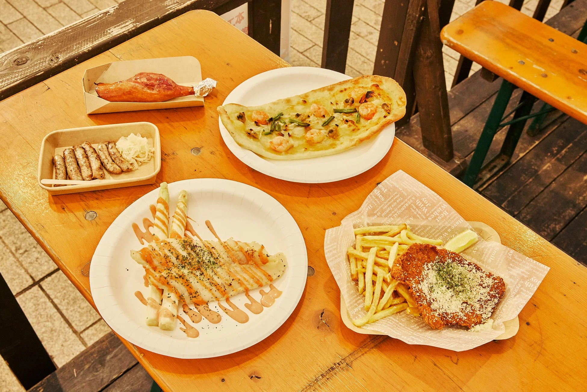 木製のテーブルに、ローストポーク、ソーセージ、シュニッツェル、フライドポテト、エビと野菜のフラットブレッド、ホワイトアスパラガスなど、多彩な料理が並んでいます