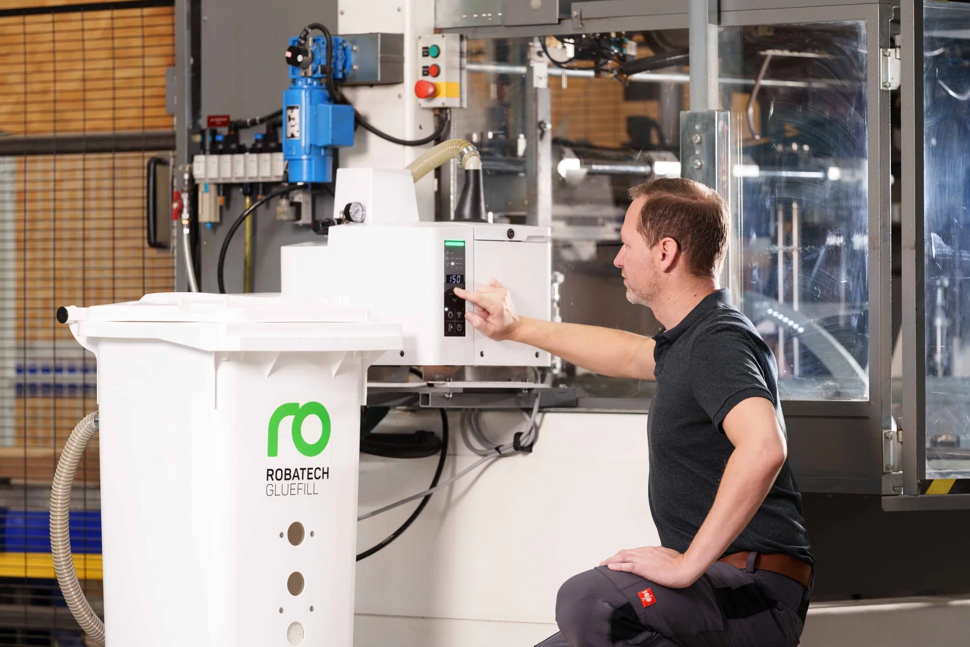 男性が産業機械「ROBATECH GLUEFILL」の操作パネルを調整している様子。接着剤充填システムが稼働する工場環境の一コマ。