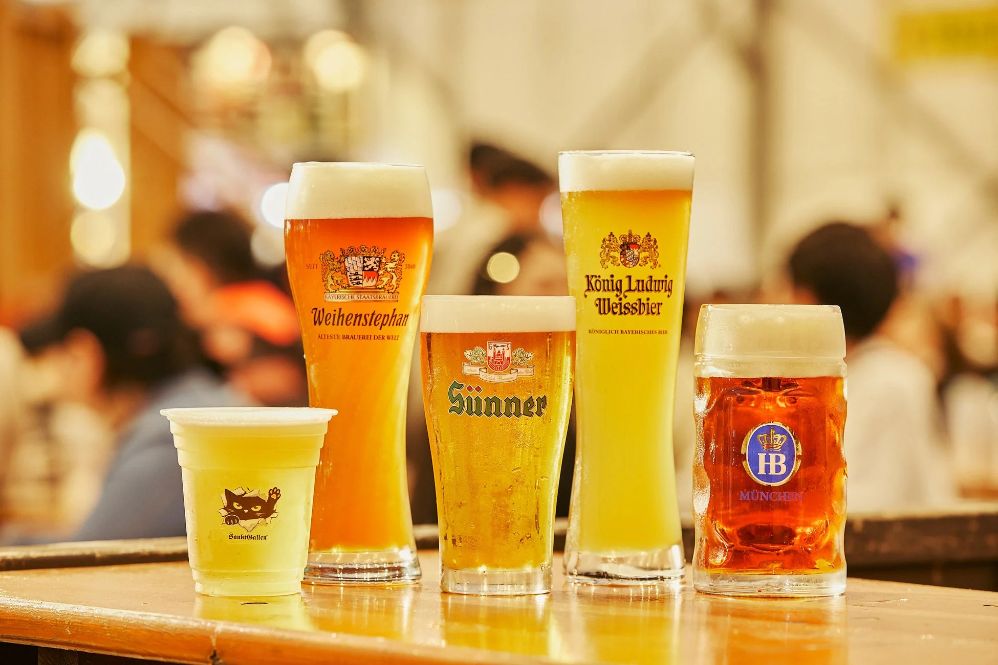 様々な種類のビールグラスがカウンターに並び、背後には賑やかなイベント会場が広がる様子