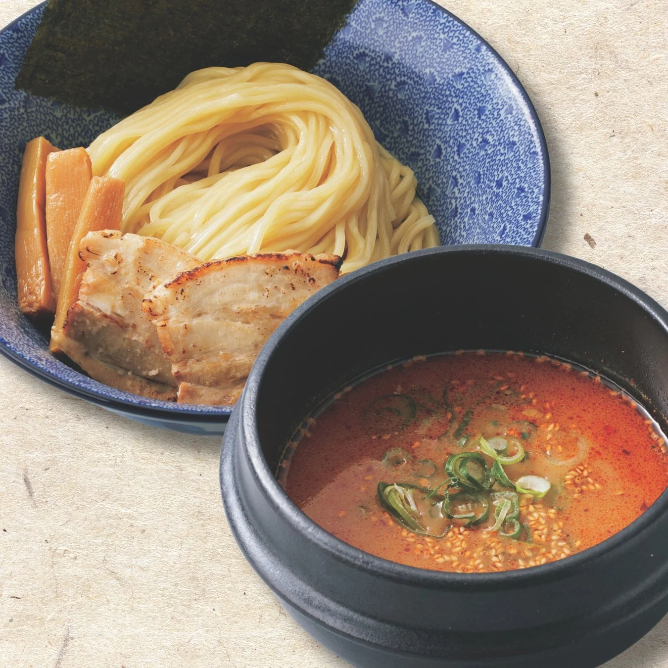 麺、チャーシュー、メンマ、海苔が盛られた器と、ネギとごまが浮いた赤く辛そうなつけ汁が別の器に用意されたつけ麺の写真です。