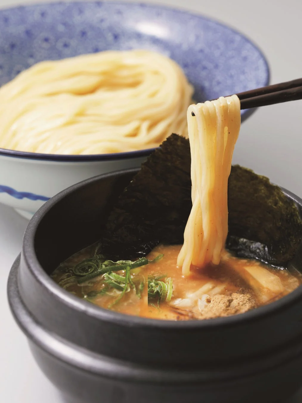 箸でつけ汁に麺を浸している瞬間を捉えたつけ麺のクローズアップ写真。食欲をそそる濃厚なつけ汁と、整然と盛られた麺が特徴的で、海苔やネギが添えられています。