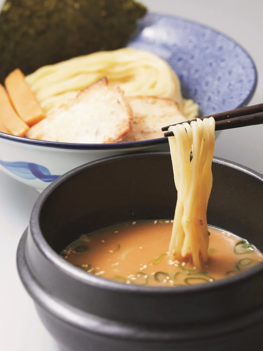 箸で麺をつけ汁に浸しているつけ麺のクローズアップ写真。麺の入った器にはチャーシューや海苔、メンマが添えられており、食欲をそそる一品です。