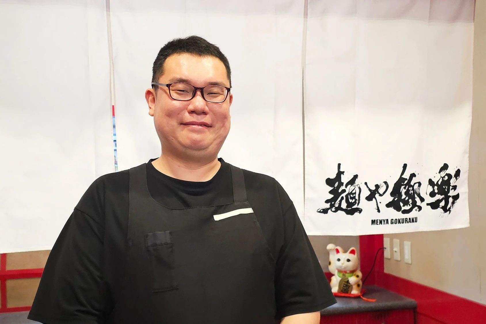 「麺や極楽」ののれんを背景に、黒いエプロンを着た男性が穏やかな笑顔で立っています。