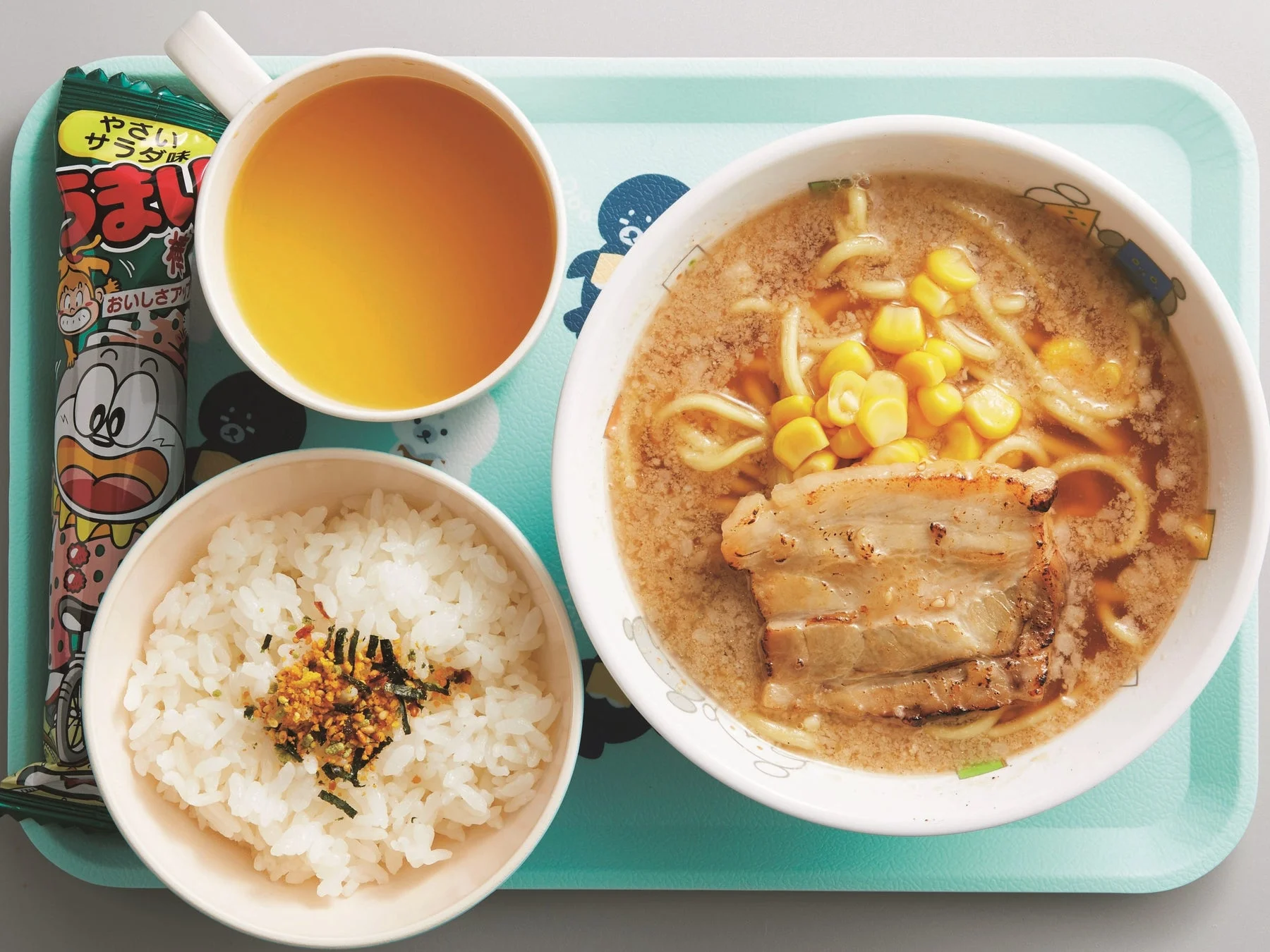 水色のトレイに置かれた日本の食事セットです。チャーシューとコーンが乗ったラーメン、ふりかけがけご飯、オレンジ色の飲み物、そしてうまい棒が並べられており、カジュアルな食卓の様子を捉えています。