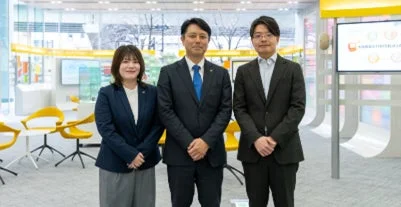 現代的なオフィス空間で、ビジネススーツを着用した男女3人が並んで立っている集合写真です。背景には黄色の椅子やテーブル、情報ディスプレイが見えます。