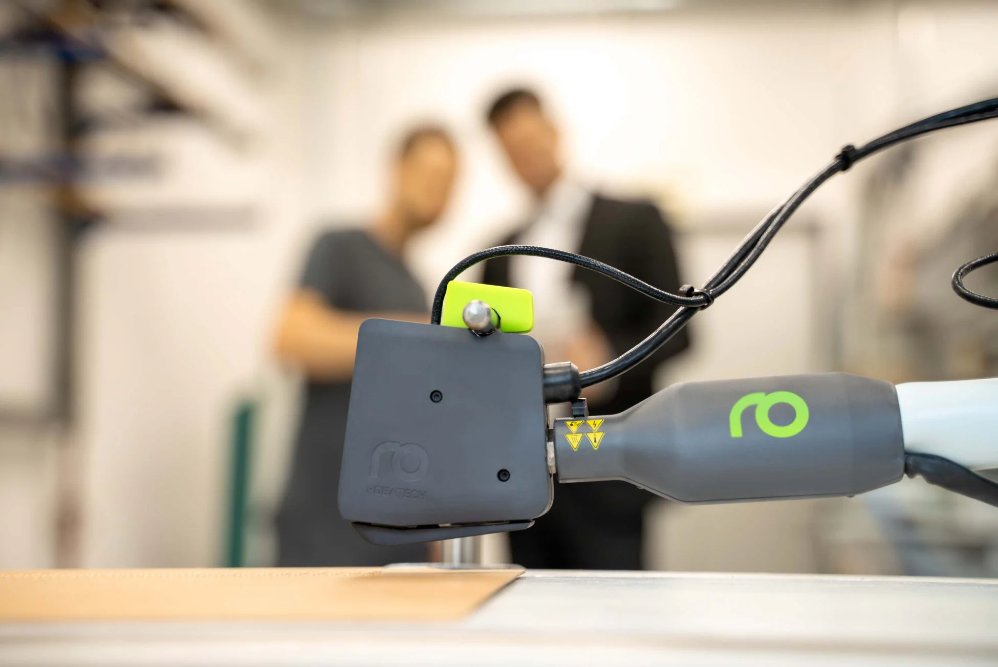 ロボットアームの先端部分がクローズアップされており、その下には素材が置かれている。背景にはぼかされた2人の人物が見える。工業用ロボットによる精密作業の様子を捉えた画像です。