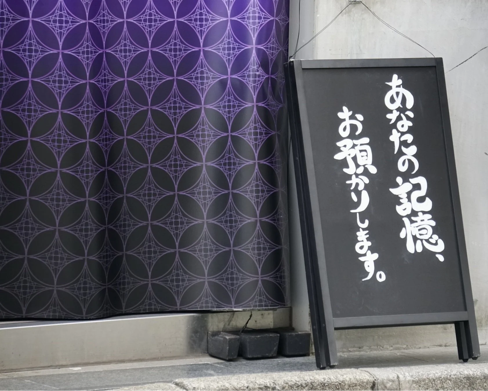 紫色の七宝文様が特徴的な店舗の入り口と、白字で「あなたの記憶、お預かりします。」と書かれた黒いA型看板が写っています。神秘的な雰囲気を醸し出しています。