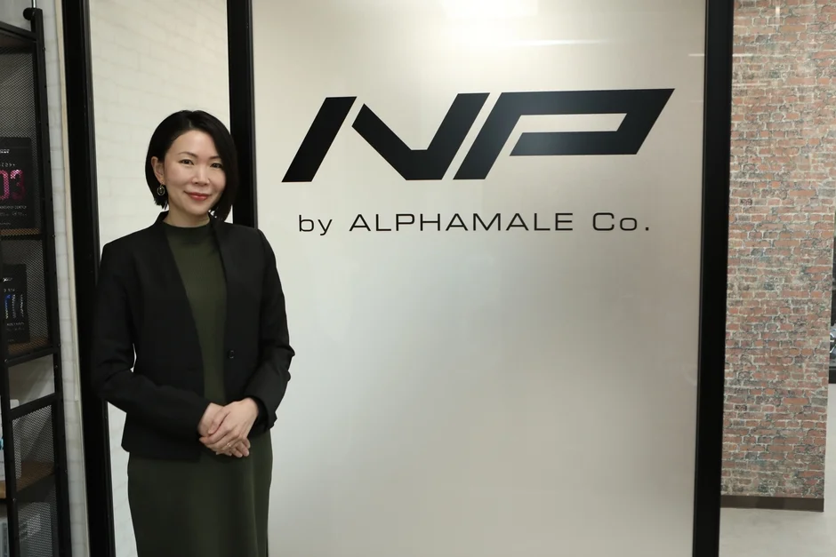 ビジネスウェアを着たアジア人女性が、企業のロゴ「NVP by ALPHAMALE Co.」が描かれたガラスの壁の前で微笑んでいます