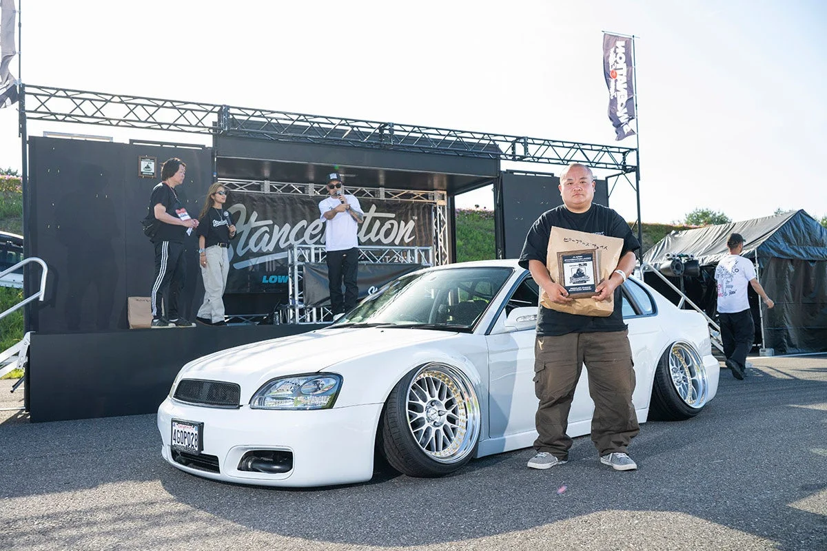 カスタムカーイベントで、極限までローダウンされた白いスバル車とアワードを持つ男性が写っています