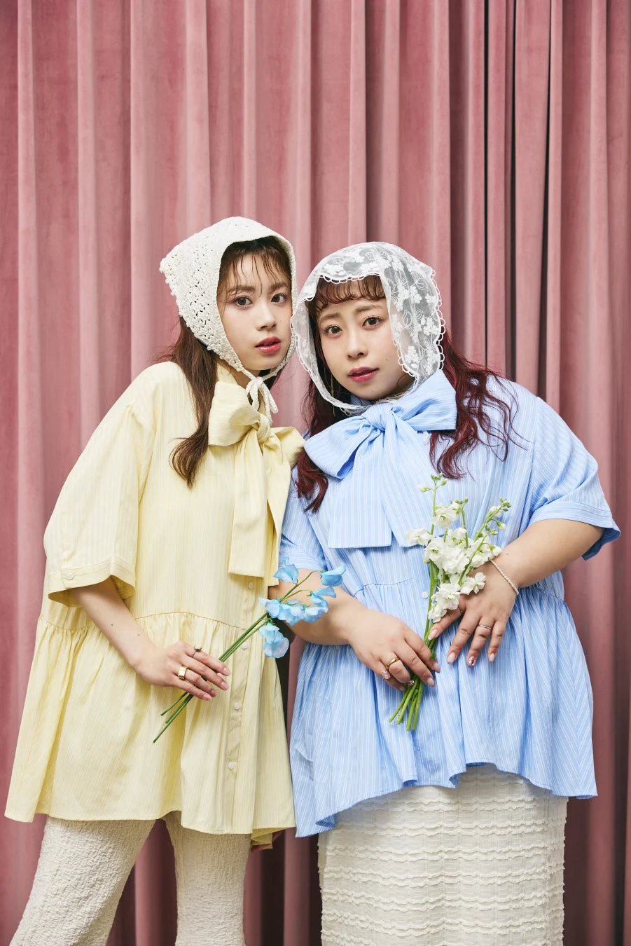 ピンクのカーテンを背景に、黄色と水色のパステルカラーの衣装を身につけた2人の女性が写っています。それぞれレースのボンネットをかぶり、花束を持って優しい表情をしています。可愛らしく、ドーリーな雰囲気のファッションポートレートです。