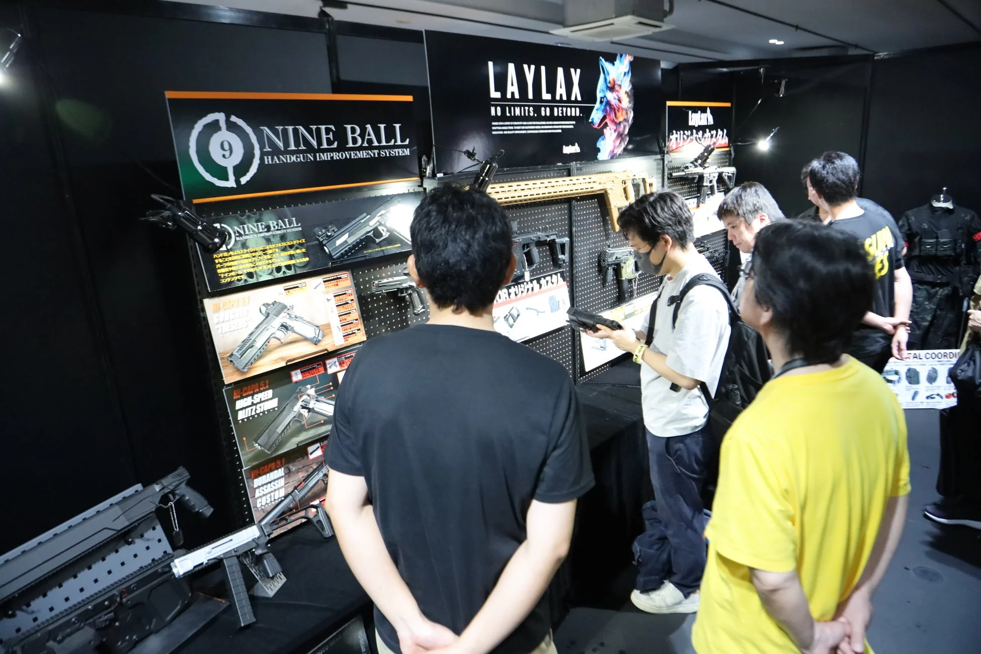 展示会または店舗のブースで、複数の男性がエアガンやカスタムパーツのディスプレイを見ている様子