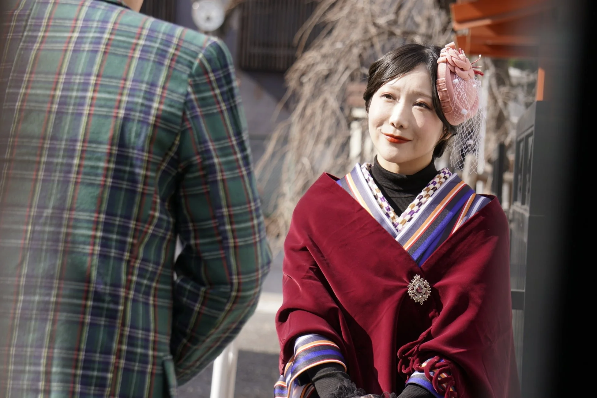鮮やかな赤色のベルベットのショールと個性的なヘアアクセサリーを身につけた女性が、屋外で微笑んでいます。伝統的な装いにモダンな要素が融合した、上品なファッションが印象的で、卒業式などのフォーマルな場面を思わせます。