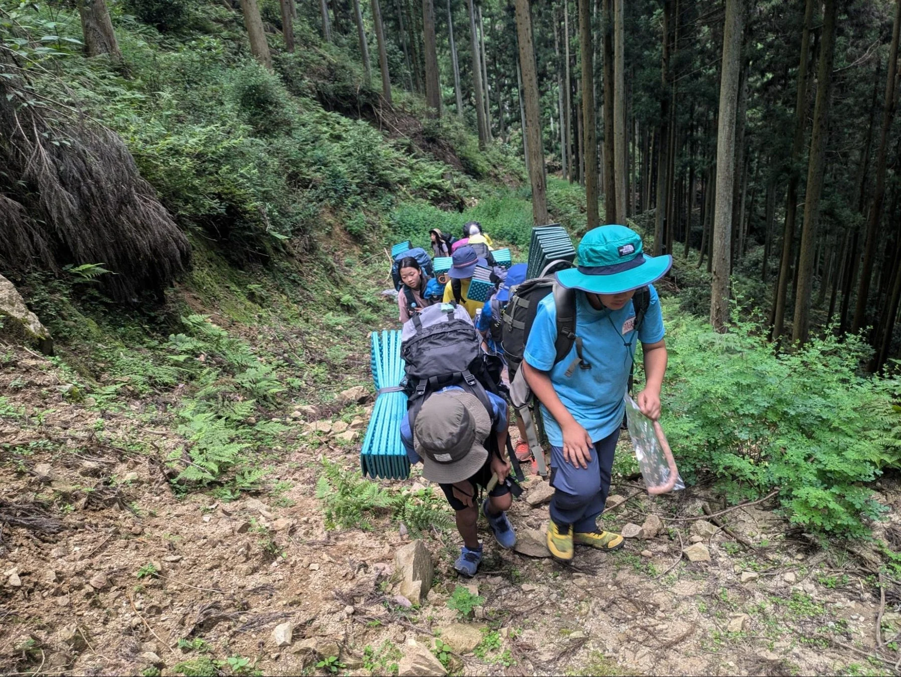 大きなリュックと寝具を背負って山道を歩く子供たちのグループ