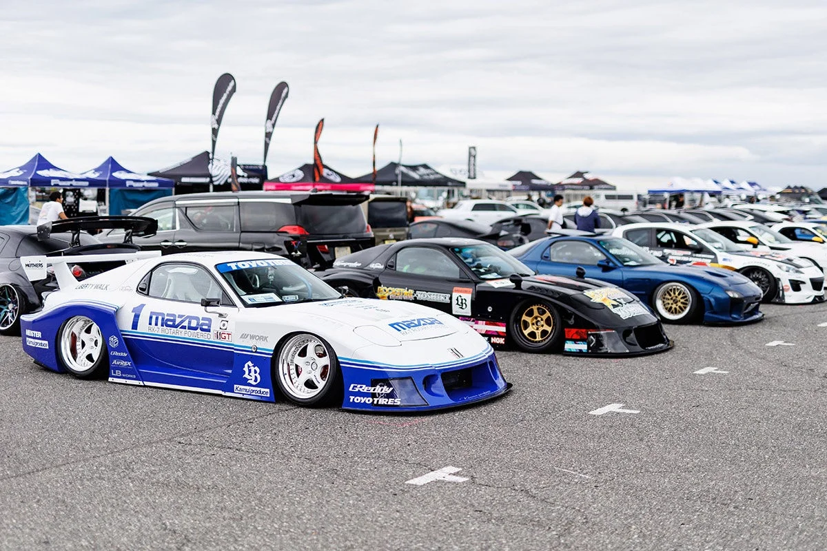 屋外のイベント会場に並べられた、白と青の目立つRX-7を先頭とする複数のカスタムカーの様子