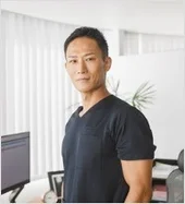 黒いシャツを着た真面目な表情のアジア人男性が、オフィスのような場所で立っているポートレート