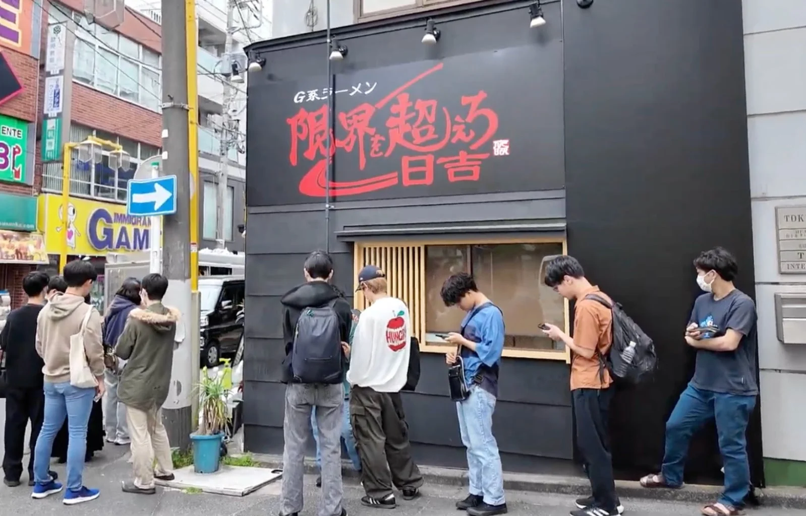 日本の街角で、多くの若者が「G系ラーメン 限界を突破 日吉」というラーメン店の前に列をなして待っている様子
