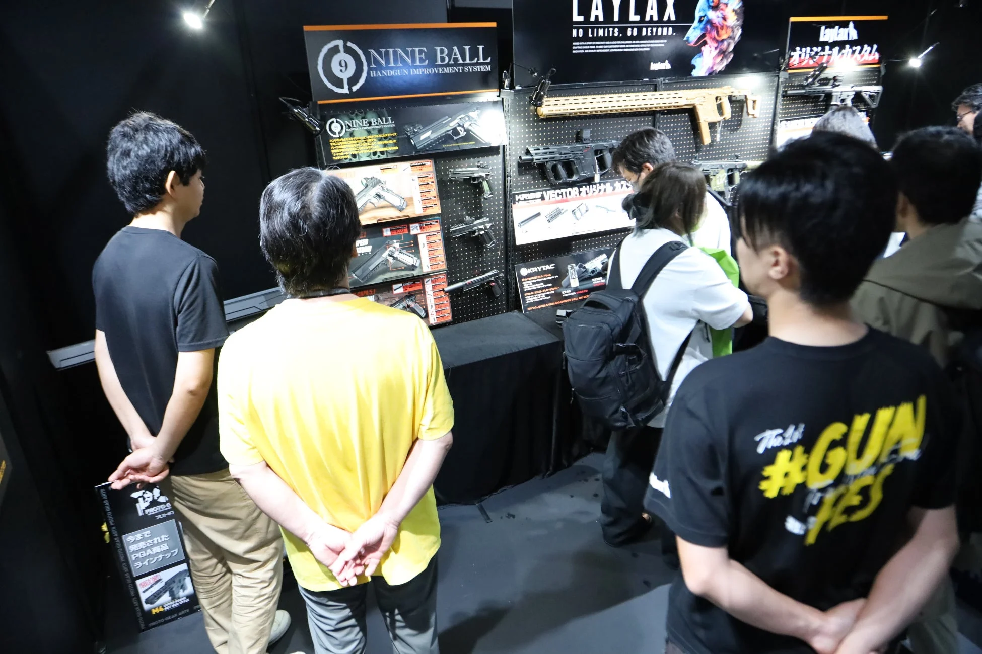 複数の人々がエアガンや関連アクセサリーの展示ブースを見ている様子