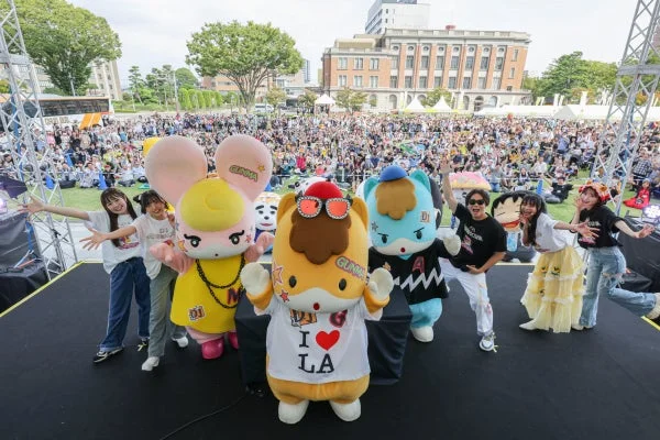 屋外イベントのステージで、複数のマスコットキャラクターとパフォーマーたちが観客を前にポーズ