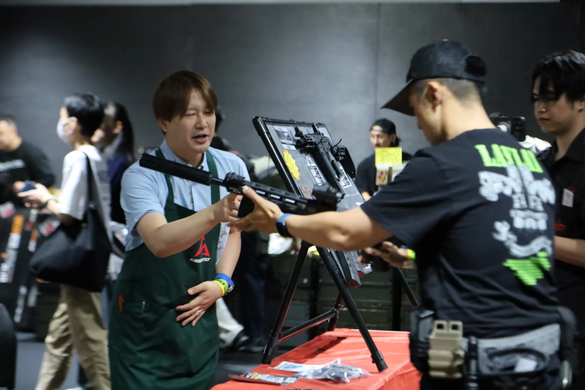 エアソフトガンのイベント会場で、エプロンを着用したスタッフが来場者に対して銃器のレプリカを説明しています