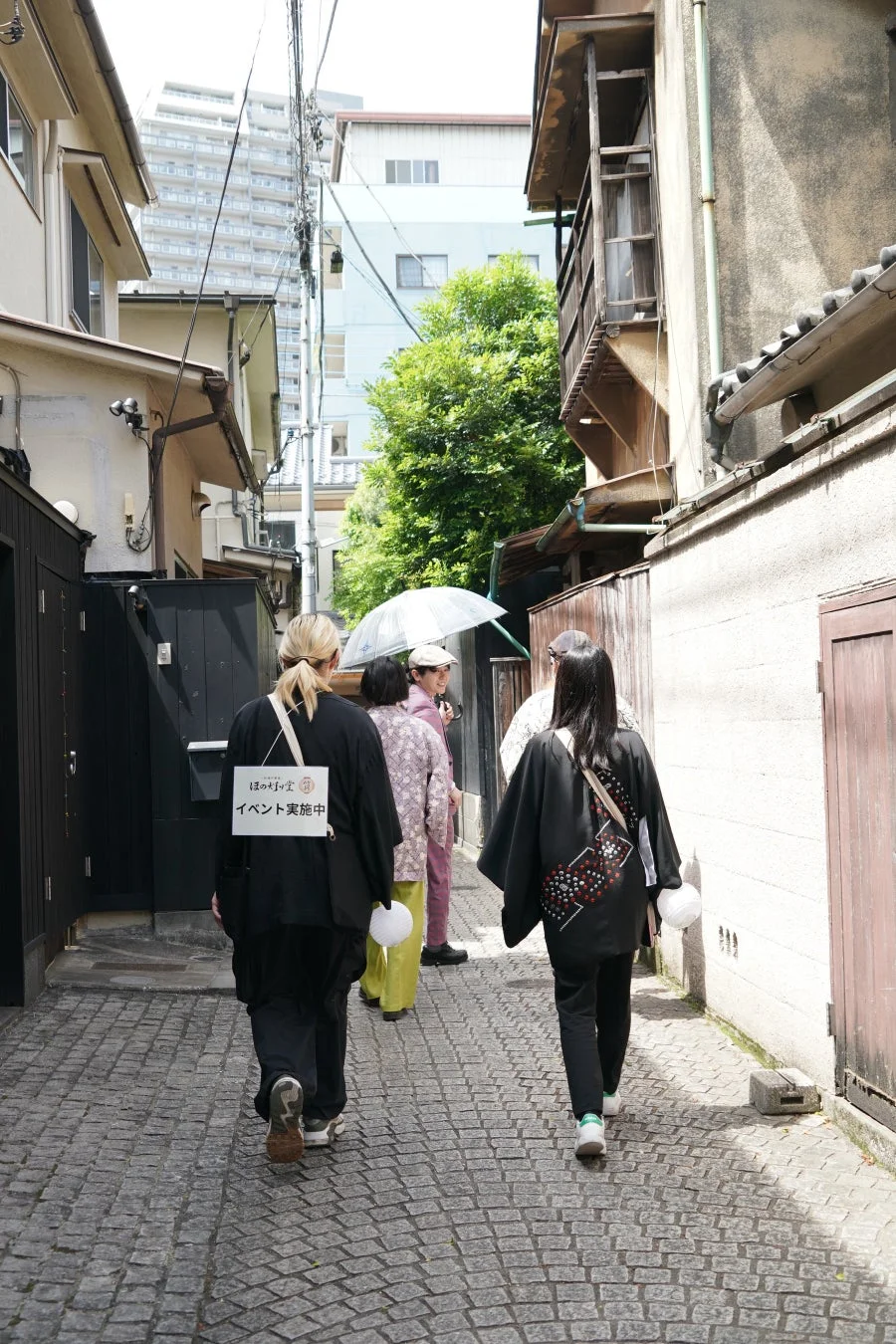 石畳の狭い路地を歩く人々が捉えられています。一人の背中には「ほのめき堂 イベント実施中」と書かれた看板があり、何らかのイベントが行われている様子です。周囲は伝統的な建物と現代的なビルが混在する日本の街並みで、夏の日の散策の雰囲気が感じられます。