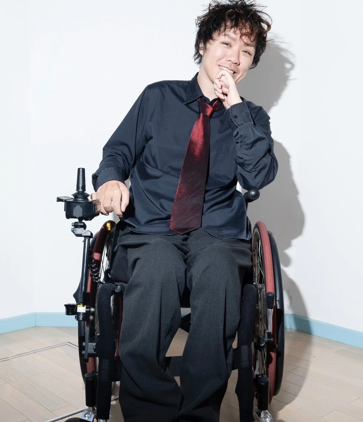 電動車椅子に座った男性が、黒いシャツと赤いネクタイを着用し、カメラに向かって笑顔を見せています