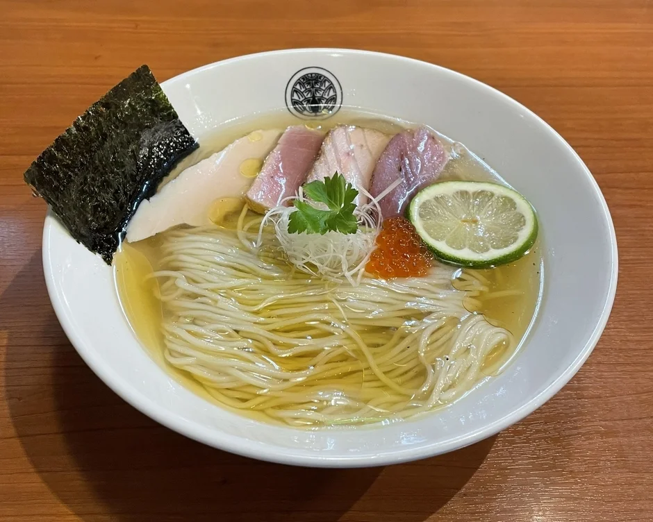 木目調のテーブルに置かれた、透き通ったスープのラーメン。細麺の上に、鶏肉とピンク色のチャーシュー、ライム、海苔、イクラ、薬味が美しく盛り付けられており、あっさりとした上品な一杯に見えます。