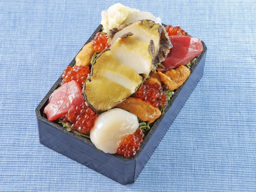 豪華な海鮮丼が黒い弁当箱に盛り付けられています。アワビ、マグロ、イクラ、ウニ、ホタテなど新鮮な海の幸が色鮮やかに配置され、食欲をそそる一品です。