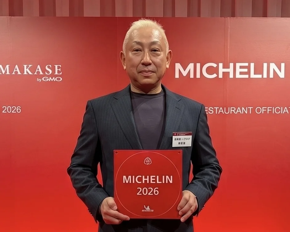 ミシュランのロゴが入った赤い背景の前で、男性が「MICHELIN 2026」と書かれた赤いプレートを持っている。ミシュラン関連のイベントで表彰されている様子。