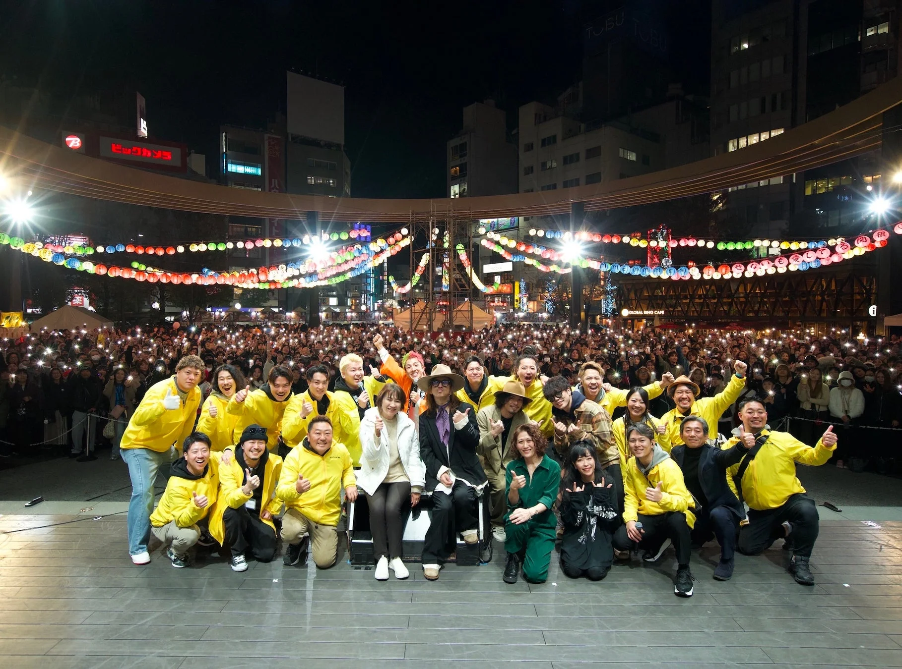 夜の広場で開催されたイベントで、ステージに立つ出演者たちが観客をバックに集合写真を撮っている様子