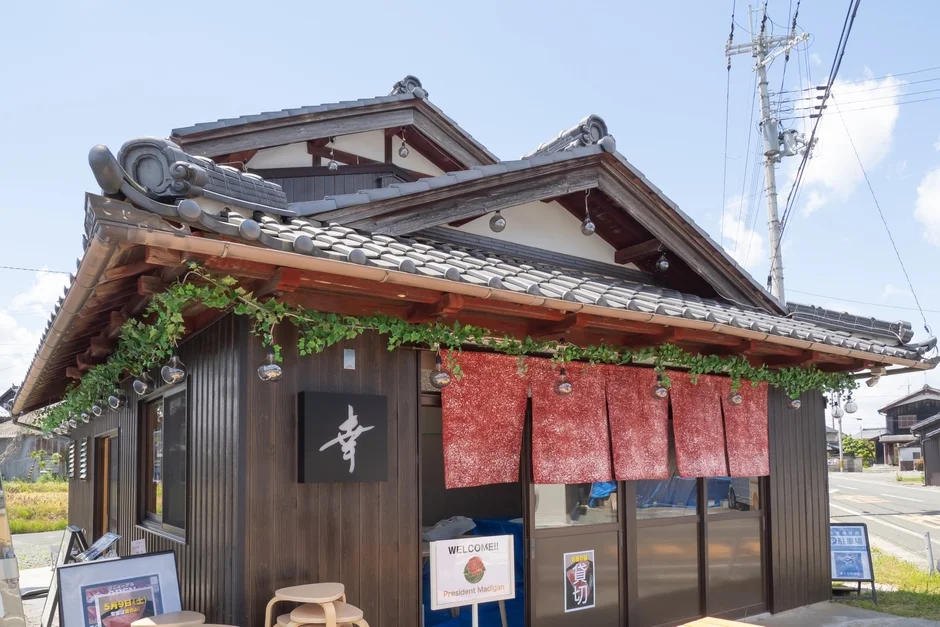 晴れた青空の下、和風建築の店舗が佇んでいます。木造の建物には「幸」の看板と赤い暖簾がかかり、「貸切」の表示も見えます。屋根には緑の装飾があり、日本の地方の風景を感じさせます。