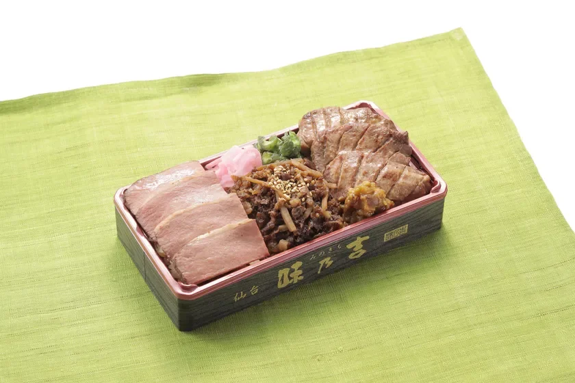 緑色の布の上に置かれた、仙台名物の牛タンを含む豪華な弁当。スライスされた肉、焼き肉、そぼろのような肉料理、漬物が入っており、彩り豊かで食欲をそそる一品です。