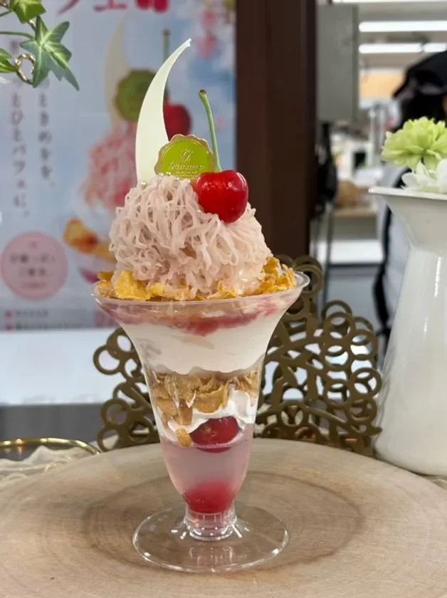 ピンク色のクリームと真っ赤なさくらんぼが目を引く、美味しそうなパフェのクローズアップ写真です。背景にはメニューらしきものが写り込んでいます。
