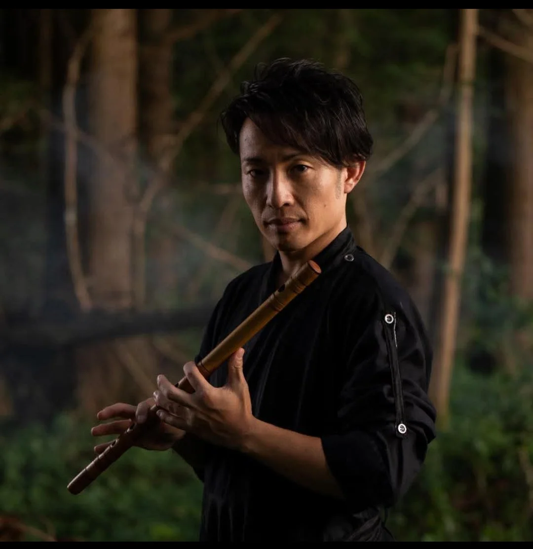森を背景に、黒い服を着た日本人男性が尺八のような和楽器を構えているポートレート