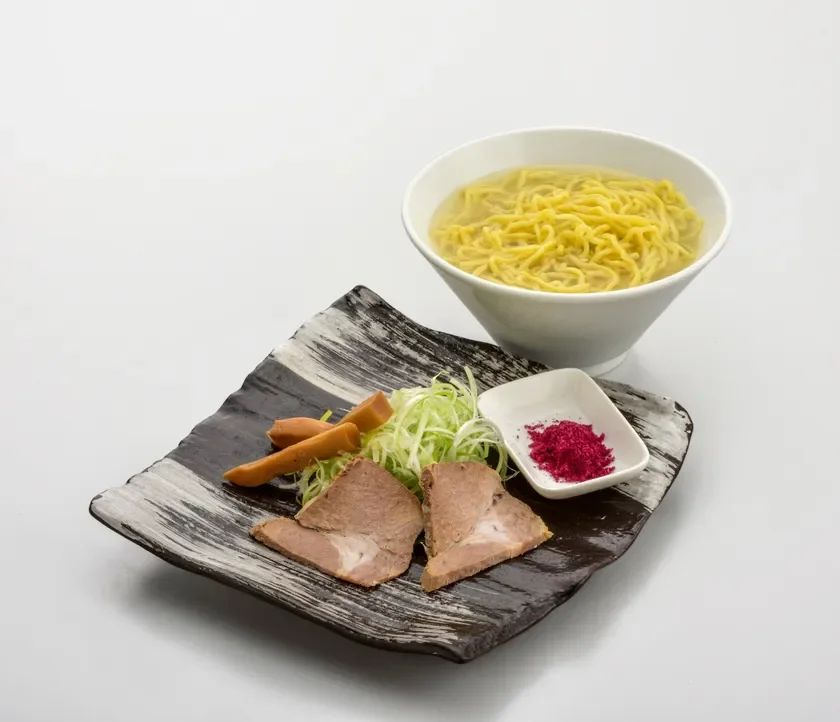 白い背景に置かれた、麺と具材が別々に盛り付けられたつけ麺のような料理です。麺は澄んだスープに入っており、具材の皿にはチャーシュー、刻みネギ、メンマ、そして赤い粉末が添えられています。