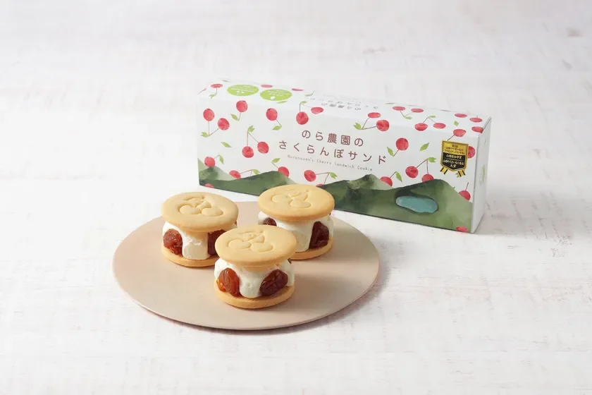 「のら農園のさくらんぼサンド」という商品が写っています。桜の絵柄が描かれた箱と、その中身と思われるサンドクッキーが3つ、お皿に盛り付けられています。クッキーは白いクリームとさくらんぼのコンポートを挟んでおり、モンドセレクション金賞受賞の表記も見られます。
